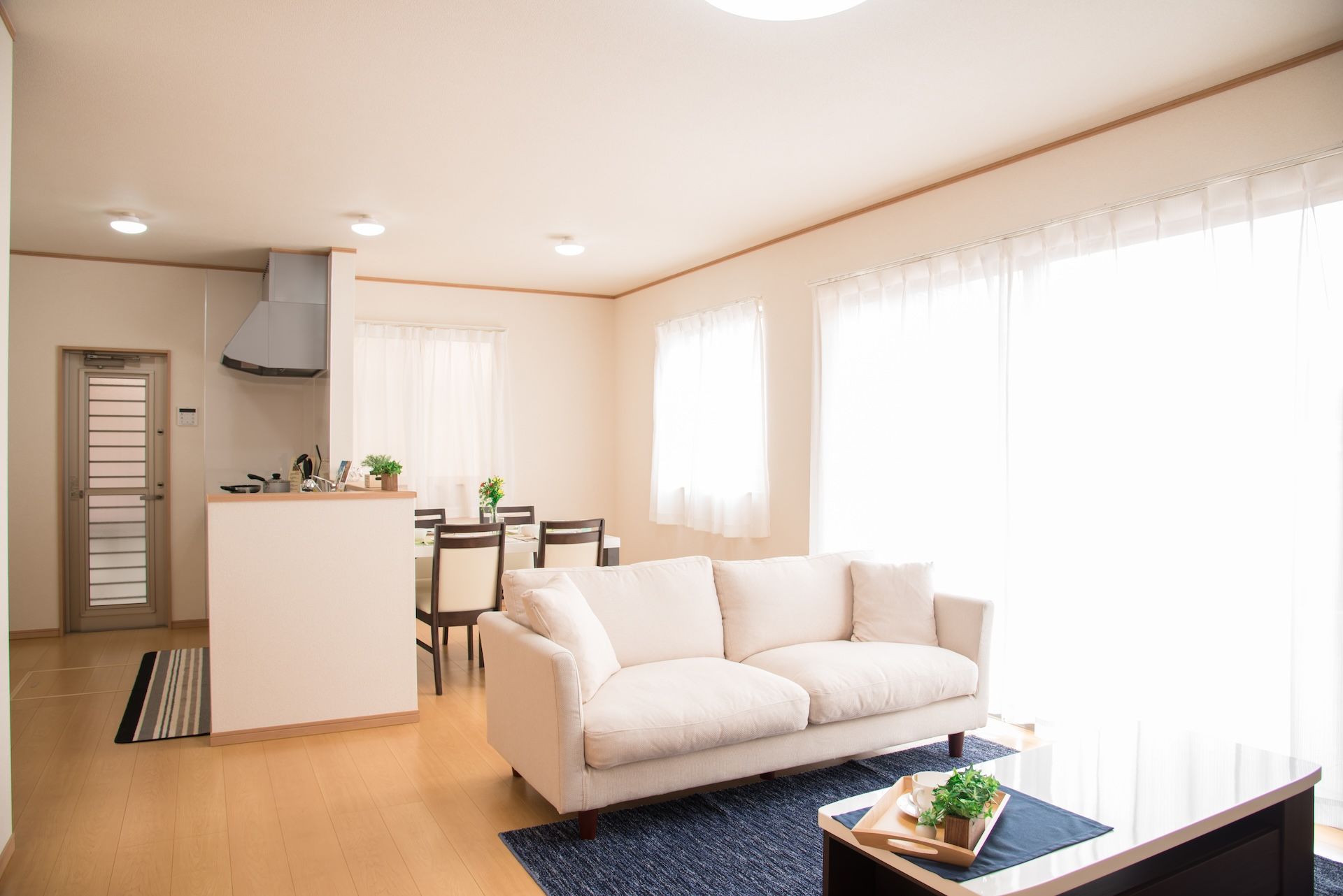 一個明亮、現代、開放式的日式公寓或房屋的客廳和用餐區。房間配有淺色木地板、米白色牆壁、輕薄的白色窗簾、一張放置在深藍色地毯上的大型米色沙發,後方可見廚房櫃檯。