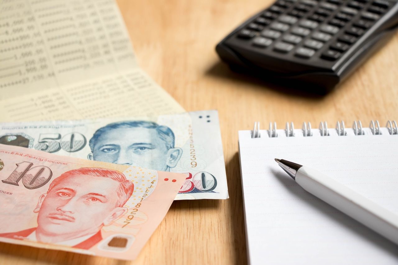 木質桌面的特寫鏡頭，桌上放著新加坡幣的十元和五十元紙鈔。紙鈔旁邊有一本白色的螺旋筆記本和一支白筆。背景處有一本模糊的銀行存摺和一台黑色的計算機，整體構圖象徵著個人理財、預算規劃或儲蓄。