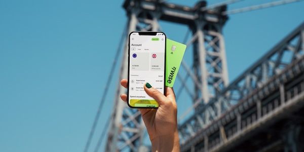 在城市大橋背景下,一手握著手機顯示 Wise App 帳戶餘額面(包含歐元與英鎊帳戶),另一手拿著亮綠色的 Wise 個人卡。